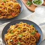 Two bowls of cheesy taco spaghetti served with a refreshing drink, garnished with chopped cilantro, and accompanied by two dipping sauces in the background.